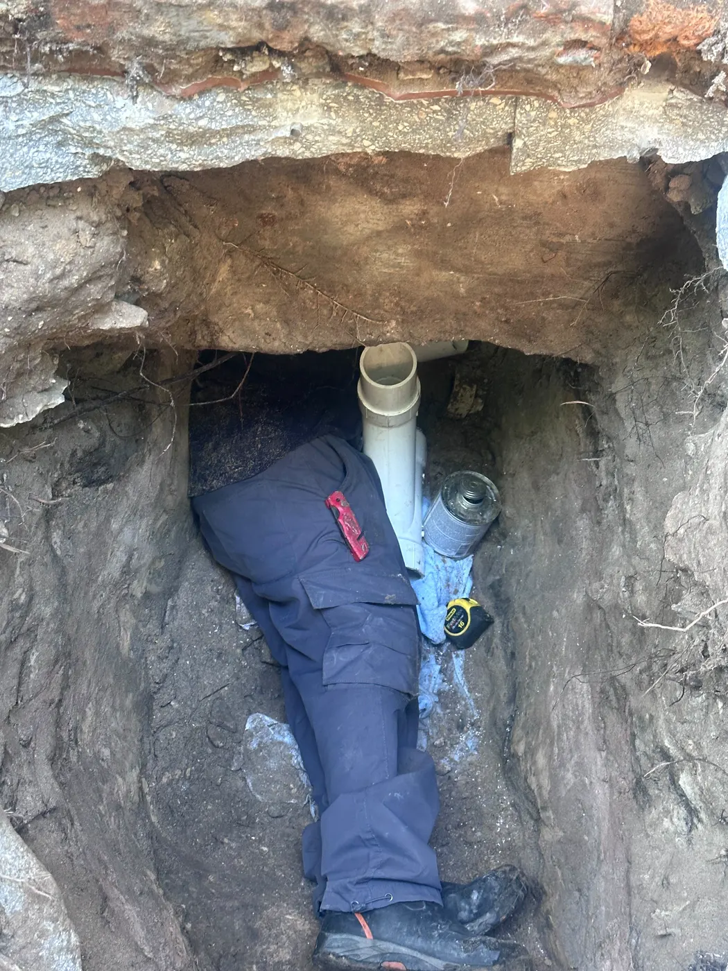 Technician lying in a trench fitting new PVC sewer pipe under a brick foundation