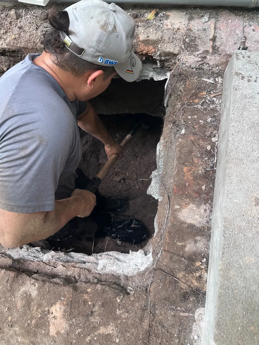 Full House technician kneeling in a hand-dug excavation beside a brick wall, working on the sewer connection