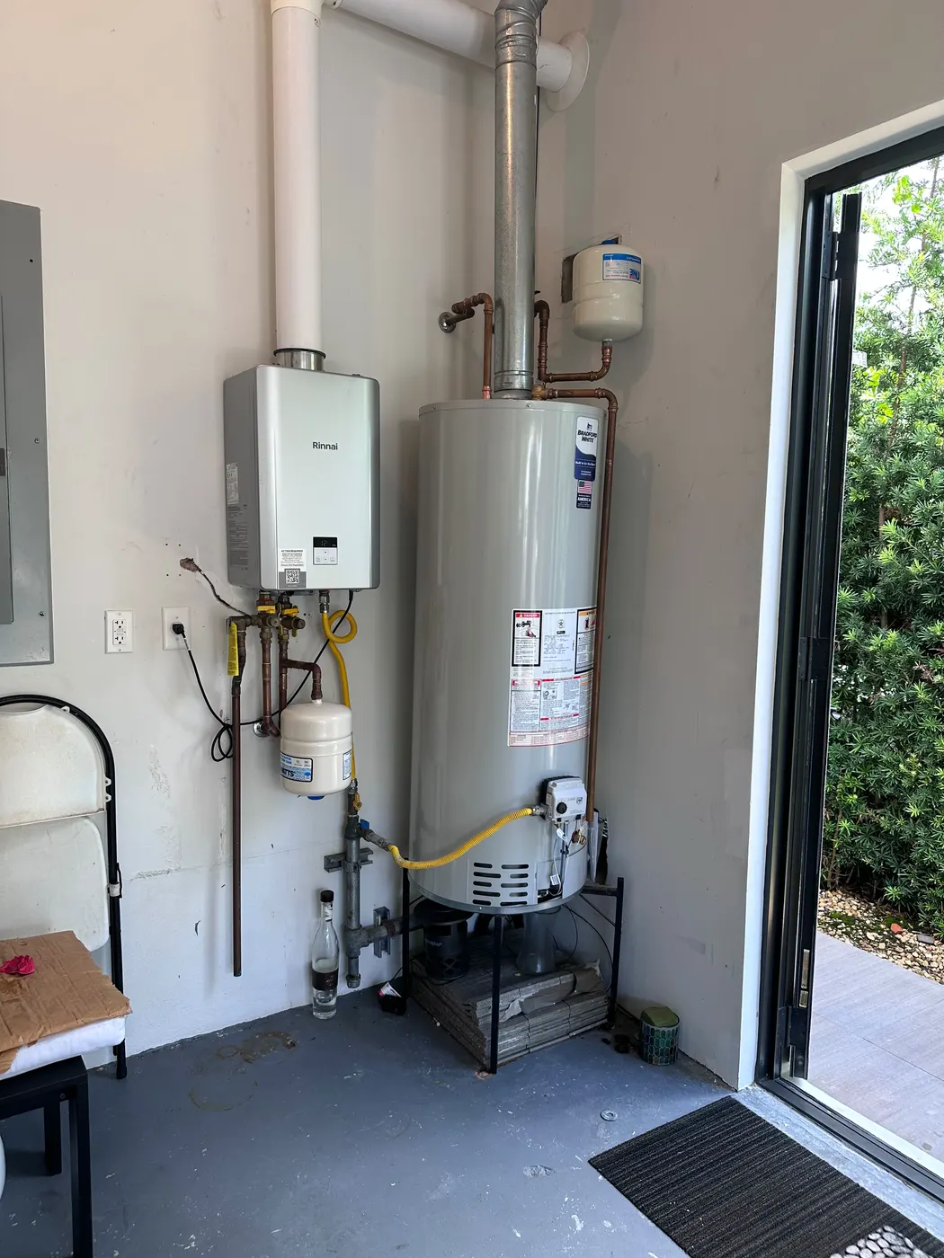 Garage utility corner with a newly installed tank water heater and tankless unit, the side door open to the garden